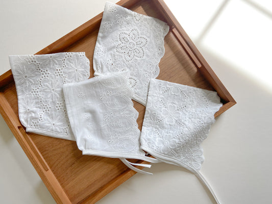 Four white bonnets are on the wooden tray. Baby bonnets are made from embroidered white cotton fabric with eyelets and have ties.  Each bonnet features embroidery in a different design with a chevron edge. One is in the form of leaves, one with clover and paisley design, one like buttercup or lotus flowers, and one like dahlia flower. 
