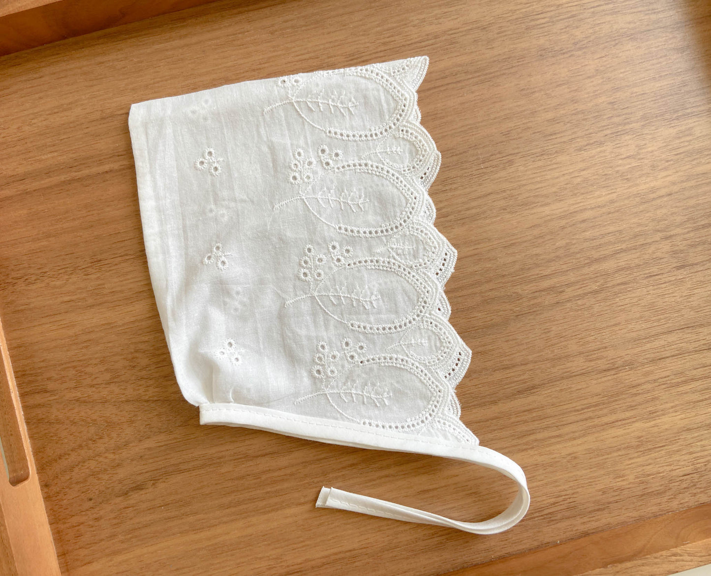 Baby bonnet is made from embroidered white cotton fabric with eyelets and has ties.  Bonnet features embroidery with leaves and a chevron edge. The leaves embroidered on the bonnet are simple, oval in shape, and have a pointy end, like apple tree, leaves. Each chevron is shaped with a smaller leave, the larger leaves are placed between the small leaves. 